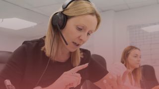 Female recruitment consultant wearing headset. She is on the phone, using her hands to communicate - one finger is pointed and the other hand is outstretched as she counts. There is a pink overlay over the image,